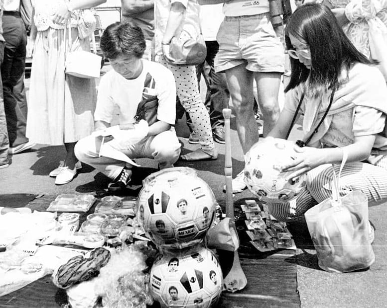 Turistas japoneses revisan mercancía mundialista; un balón con rostros de seleccionados mexicanos costó 28 mil pesos del momento. Previo a la inauguración, se supo del intento de los organizadores de impedir que 5 mil vendedores se colocaran afuera del Estadio Azteca y del Estadio Universitario, con el pretexto de garantizar la seguridad; en realidad querían rentar 50 quioscos al exterior de las sedes para vender souvenirs, sin competencia. Foto: Gildardo Solís/Archivo EL UNIVERSAL.
