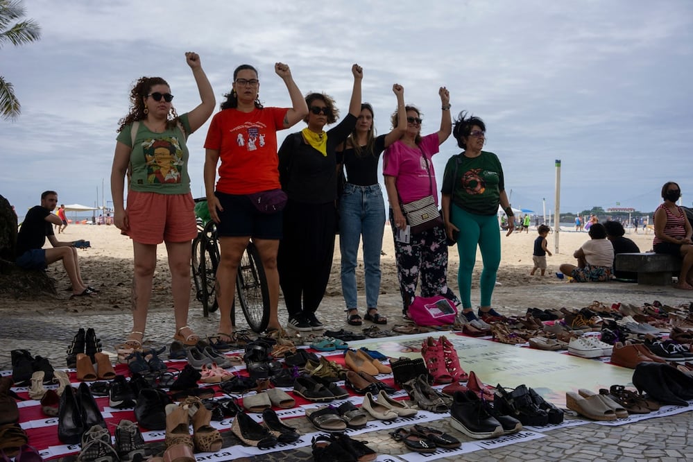 Mujeres posan frente a pares de zapatos con los nombres de mujeres víctimas de feminicidio en Brasil, durante un evento por el Día Internacional de la Eliminación de la Violencia contra la Mujer en la playa de Copacabana, Río de Janeiro, en 2022. FOTO: AFP