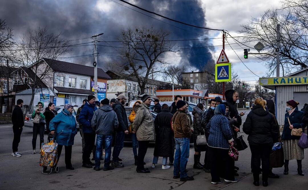La gente hace fila frente a un supermercado mientras el humo se eleva sobre la ciudad de Vasylkiv, en las afueras de Kiev, el 27 de febrero de 2022, después de que los ataques rusos durante la noche golpearan un depósito de petróleo. Foto: AFP