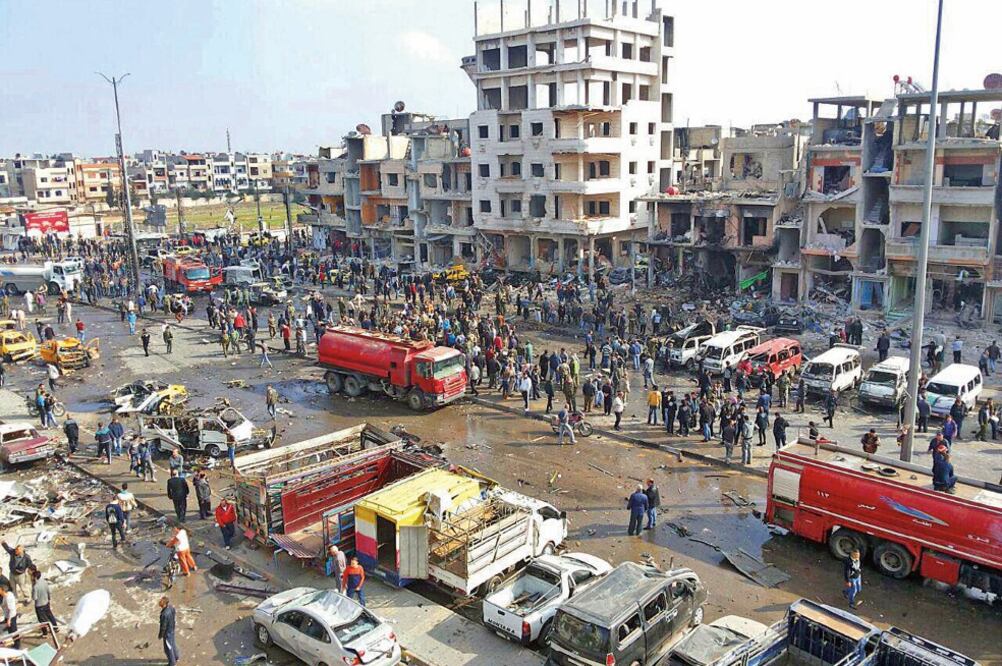 Fotografía difundida por la agencia oficial siria Sana, que muestra a decenas de personas reunidas en el sitio devastado tras dos explosiones que hubo ayer en el barrio de Zahraa, en la provincia siria de Homs (AP)