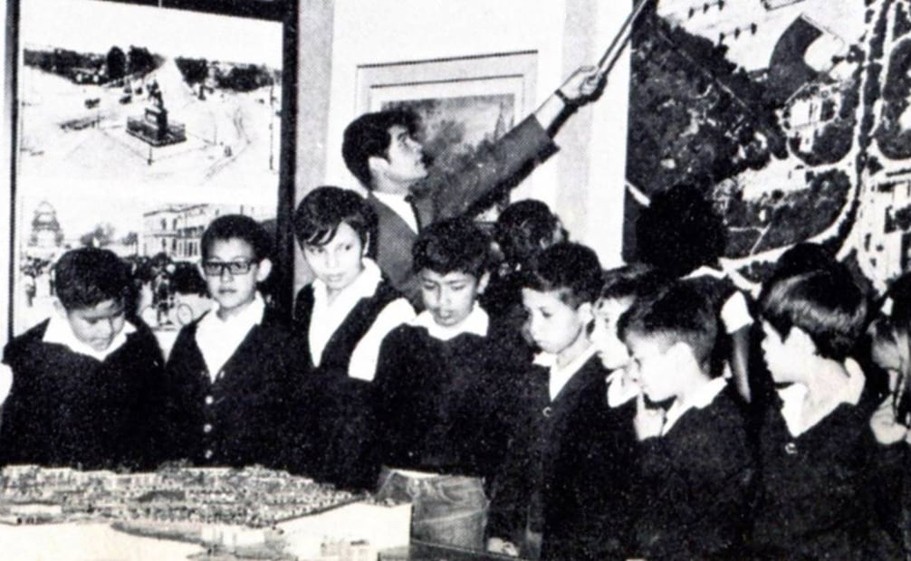 Clásica visita guiada de una escuela pública mixta para que los estudiantes aprendan acerca de urbanismo e historia en algún museo capitalino a mediados de los años setenta. Los niños observan atentos una maqueta, mientras un joven muestra a un alumno una foto aérea de algún sitio de la ciudad. Colección Villasana.