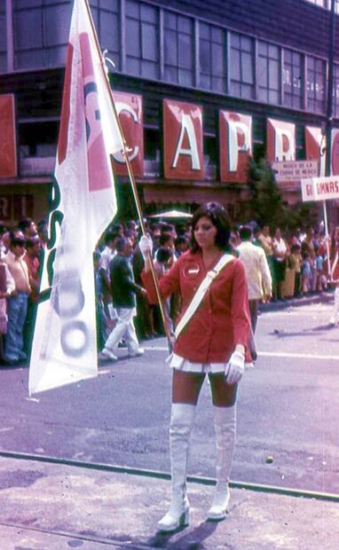 La abanderada de un grupo de jóvenes desfilando en representación de Conasupo, en la esquina de la avenida 20 de noviembre y calle República del Salvador. Además del típico conjunto de la muchacha, destaca el vistoso letrero de la zapatería CAPRI, muy famosa en décadas pasadas. Colección Villasana.