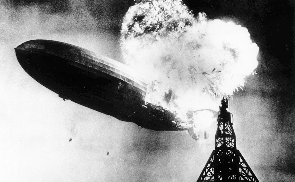 Foto de la explosión del Hindenburg. Se ve al zepelín con un incendio parcial, momentos antes de impactarse con el suelo de la base aeronaval de Lakehurst. También se ve a dos hombres, parados en la parte superior del mástil de agarre, muy cerca de las llamas. Foto: Gus Pasquarella/AP Photo