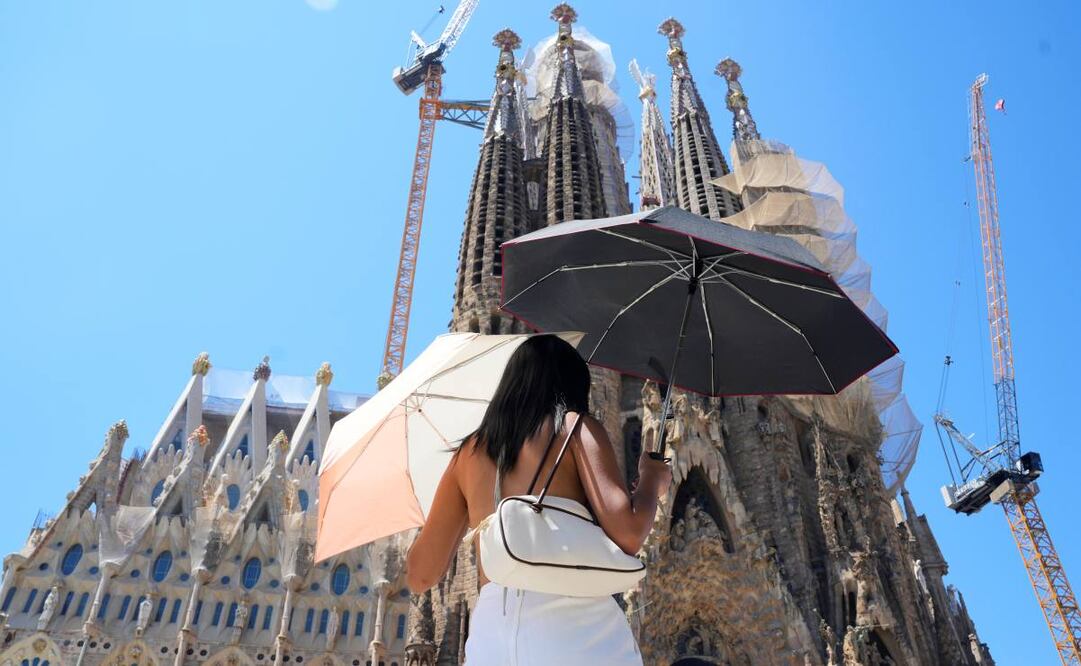 Una turista utiliza dos sombrillas para protegerse del sol frente a la Sagrada Familia en Barcelona este sábado. Catorce comunidades están este sábado en alerta por temperaturas excepcionalmente altas en el día de inicio de la primera ola de calor del verano en España. Foto: EFE