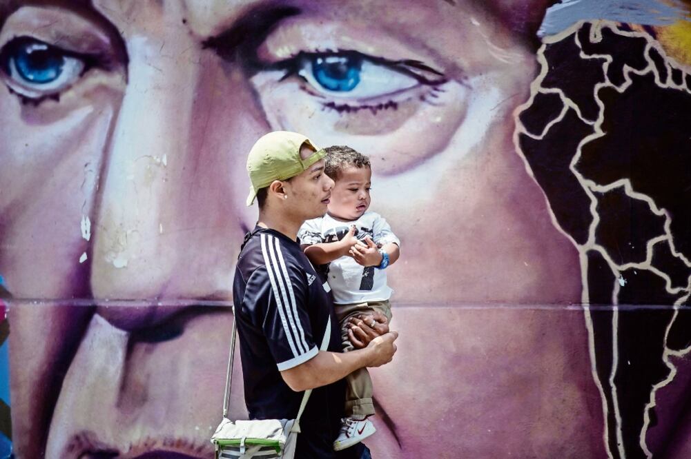 Un venezolano con un niño en brazos pasa en Caracas junto a una pared pintada con el rostro de Chávez, quien falleció de cáncer en 2013, en Cuba (MIGUEL GUTIÉRREZ. EFE)