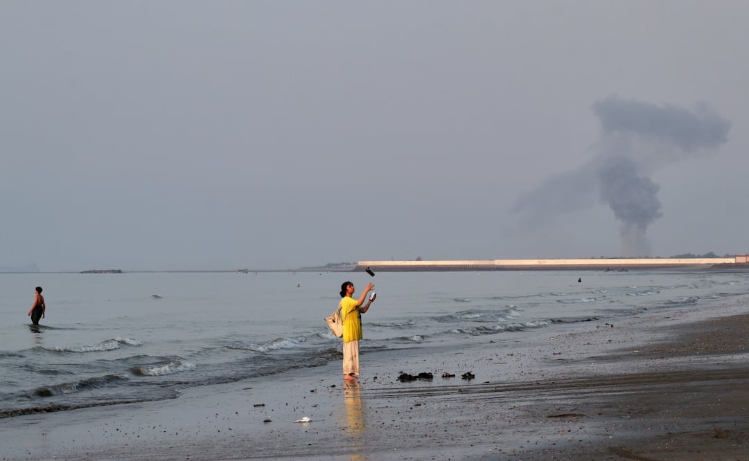 Una mujer arroja un recipiente al aire en la costa del estrecho de Ormuz, mientras a lo lejos se eleva una columna de humo tras una explosión en Irán. Foto: AP
