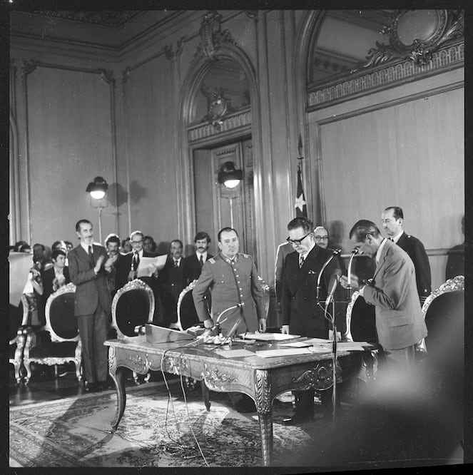 Salvador Allende leyendo un decreto, en 1972. A la derecha de éste, al fondo y en el centro de la imagen, de bigote, con pelo largo, Danilo Bartulín/ BIBLIOTECA NACIONAL DIGITAL DE CHILE, ARMINDO CARDOSO.