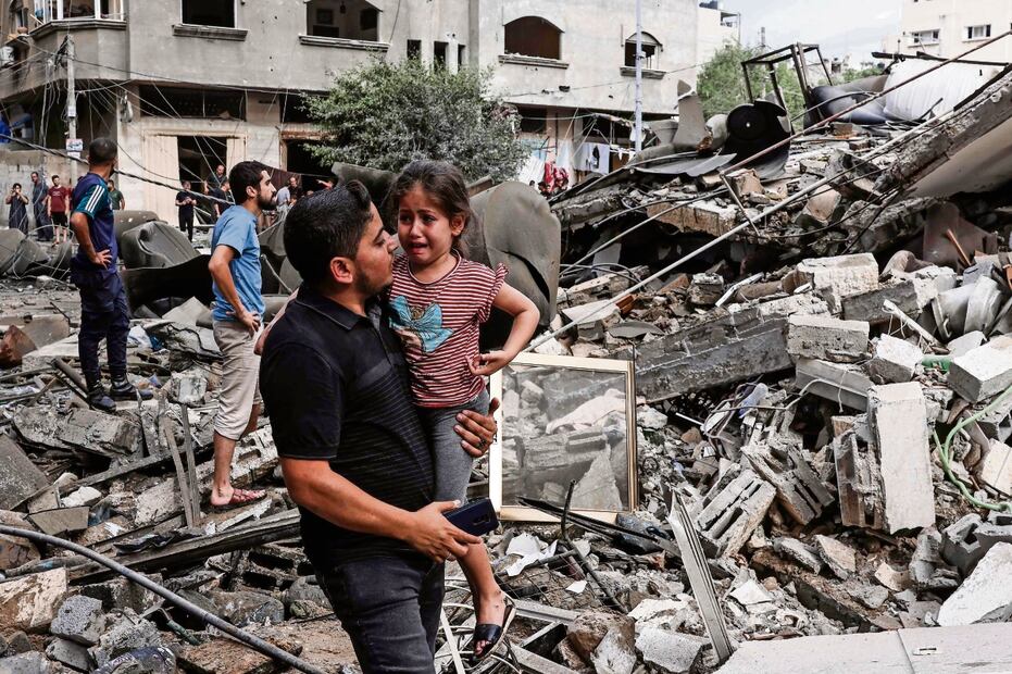 Un hombre carga a una niña llorando, mientras camina frente a un edificio destruido en un ataque aéreo israelí en la ciudad de Gaza. Foto: AFP