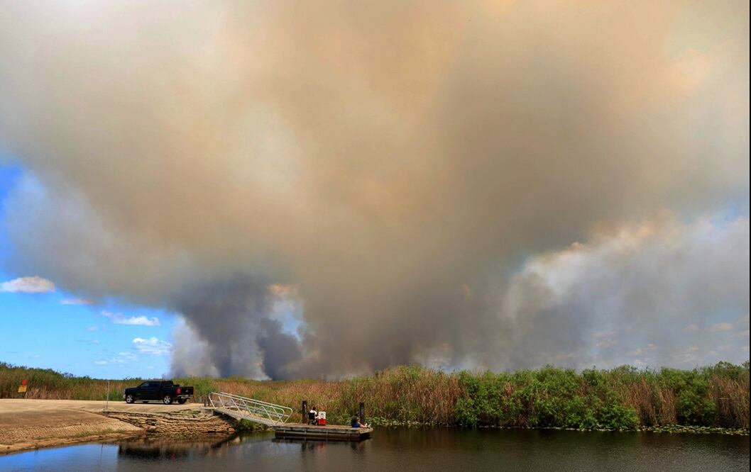Personas pescan en un canal mientras el humo se eleva tras un incendio forestal en los Everglades, Florida, el 20 de agosto de 2025. Foto: AFP