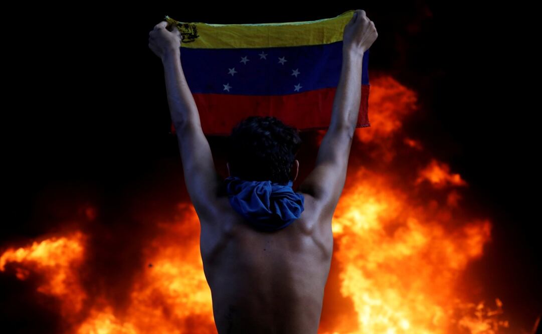 A protestor holds a national flag while standing in front of a fire burning at the entrance of a building housing the magistracy of the Supreme Court of Justice and a bank branch, during a rally against Nicolas Maduro - Photo: Carlos Garcia Rawlin/Reuters